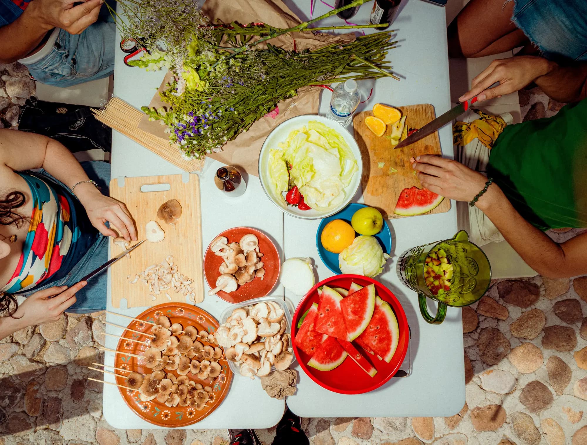 La mesa vista desde arriba: hongos, verduras, sandía y varias manos preparando la comida