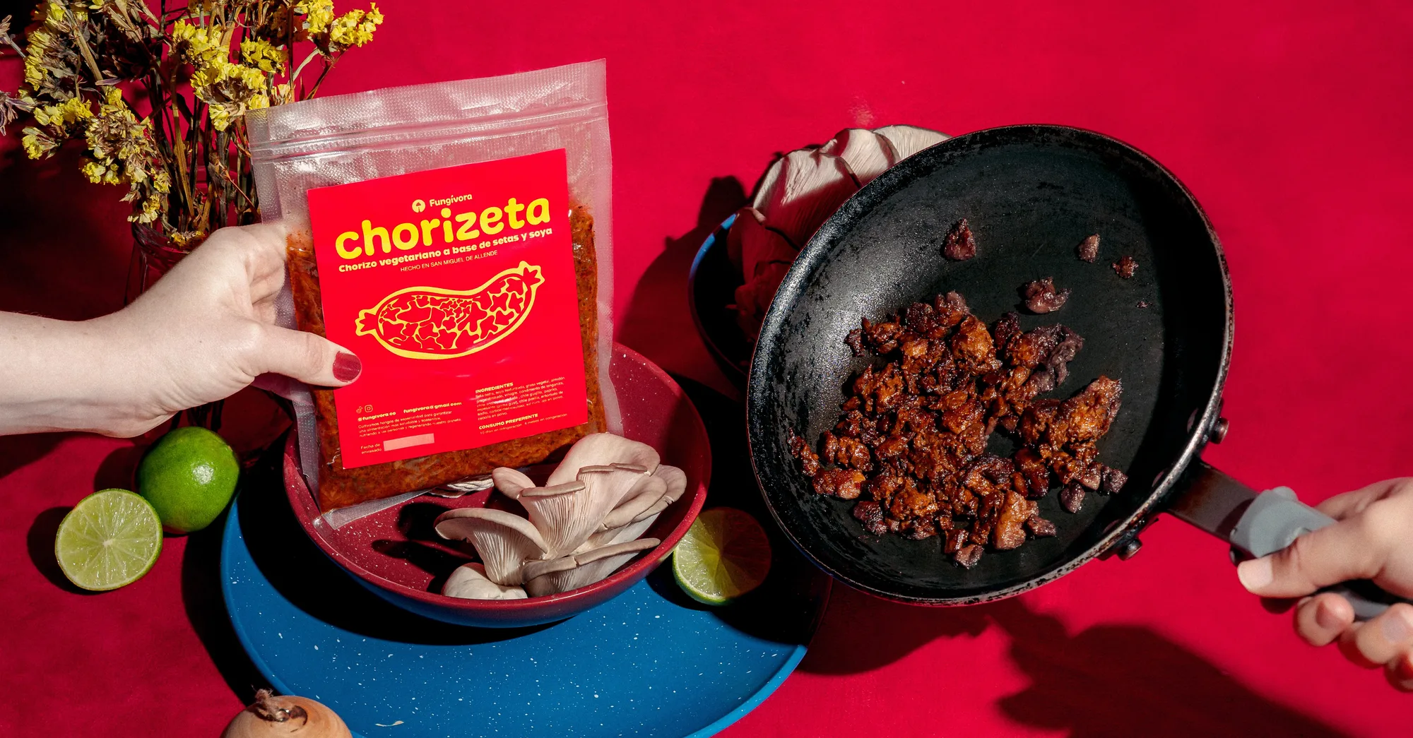 Una mano sosteniendo un paquete de Chorizeta sobre un tazón rojo de setas ostra, junto a un sartén negro con Chorizeta ya cocinada, sobre un fondo rojo