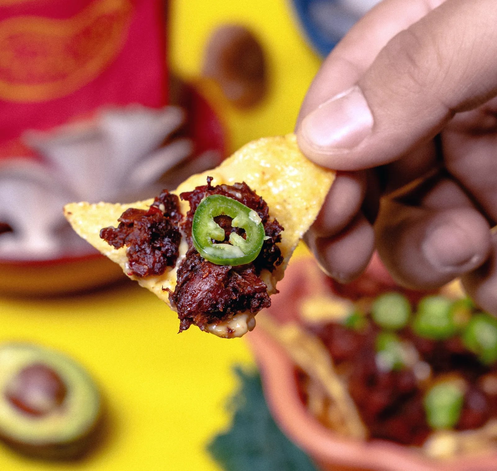 Una mano sosteniendo un nacho cargado de Chorizeta con una rodaja de jalapeño encima
